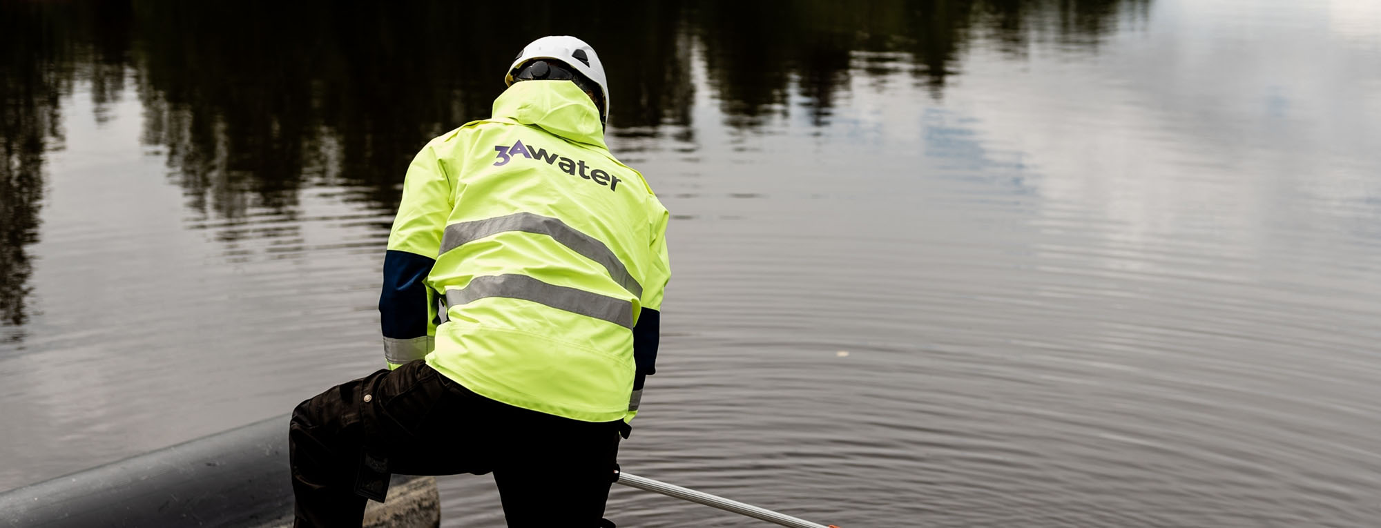 A person wearing a 3AWater branded safety jacket taking a water sample from a pond.