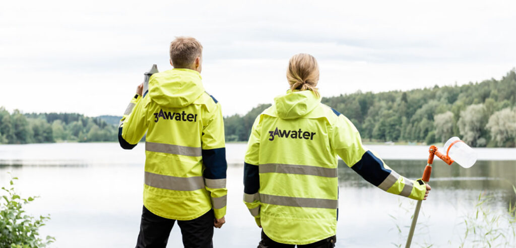 Two persons wearing 3AWater branded safety jackets in front of a lake.